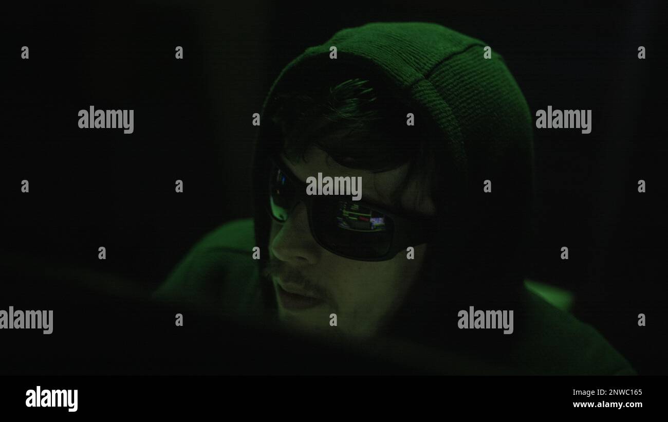 Aufnahme eines Hackers mit schwarzer Brille, der in einem dunklen Raum mit unheimlicher grüner Beleuchtung auf dem Computer tippt Stockfoto