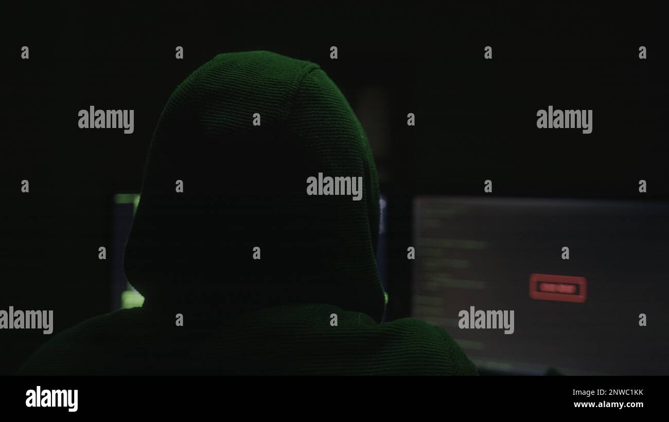 Geheimnisvoller Hacker oder Programmierer mit Hacken mit Hoodie in einem verlassenen Raum mit grünen Lichtern, Aufnahme von hinten Stockfoto