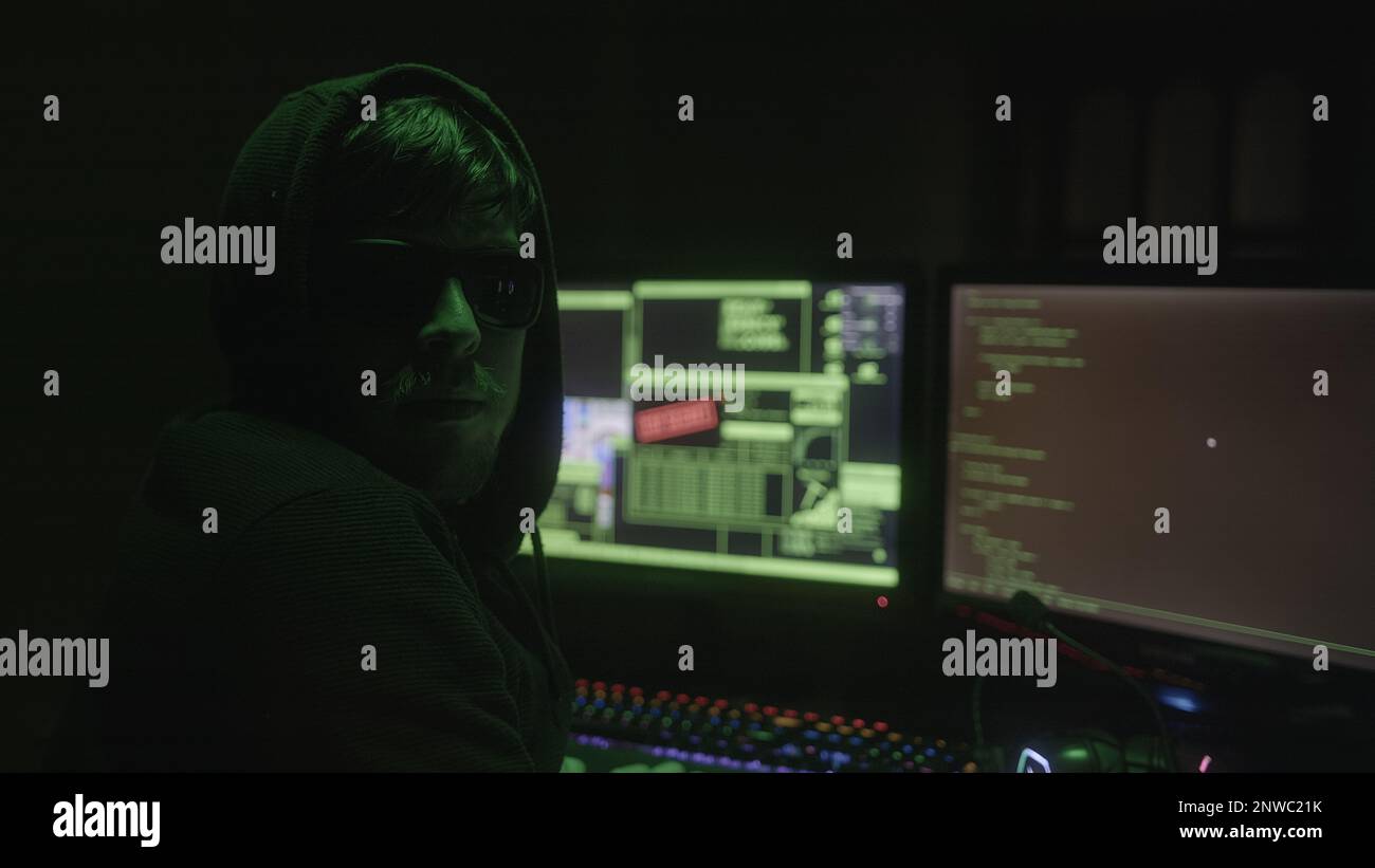 Schwarze Kapuze und Brille tragende mysteriöse Hacker oder Programmierer, die in einem verlassenen Raum mit grünen Lichtern hacken, drehen sich plötzlich um, um auf die Kamera zu schauen, Stockfoto