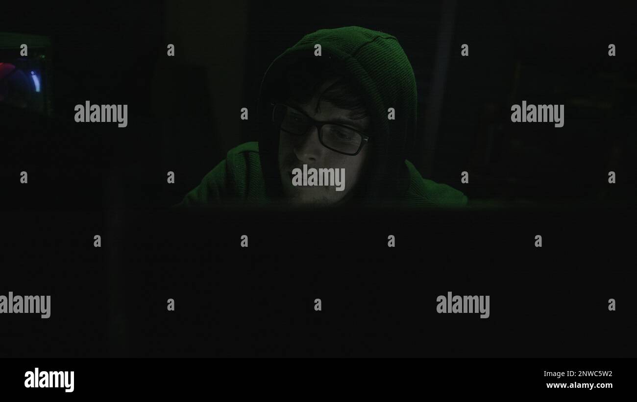 Junger Programmierer mit Hoodie und schwarzer Brille mit grüner Beleuchtung Stockfoto