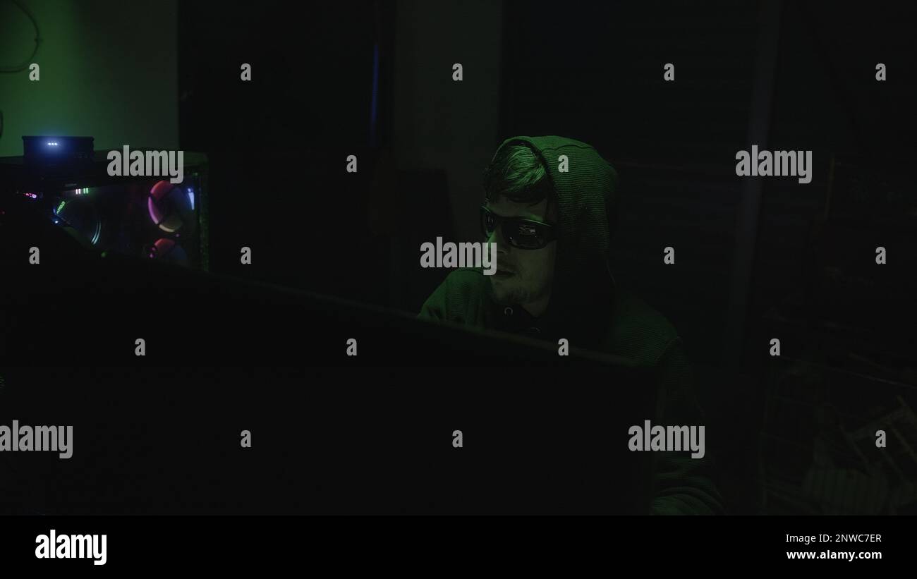 Schwarze Brille tragende Hacker-Hacking bei Computer, Green Light Ambiance Stockfoto