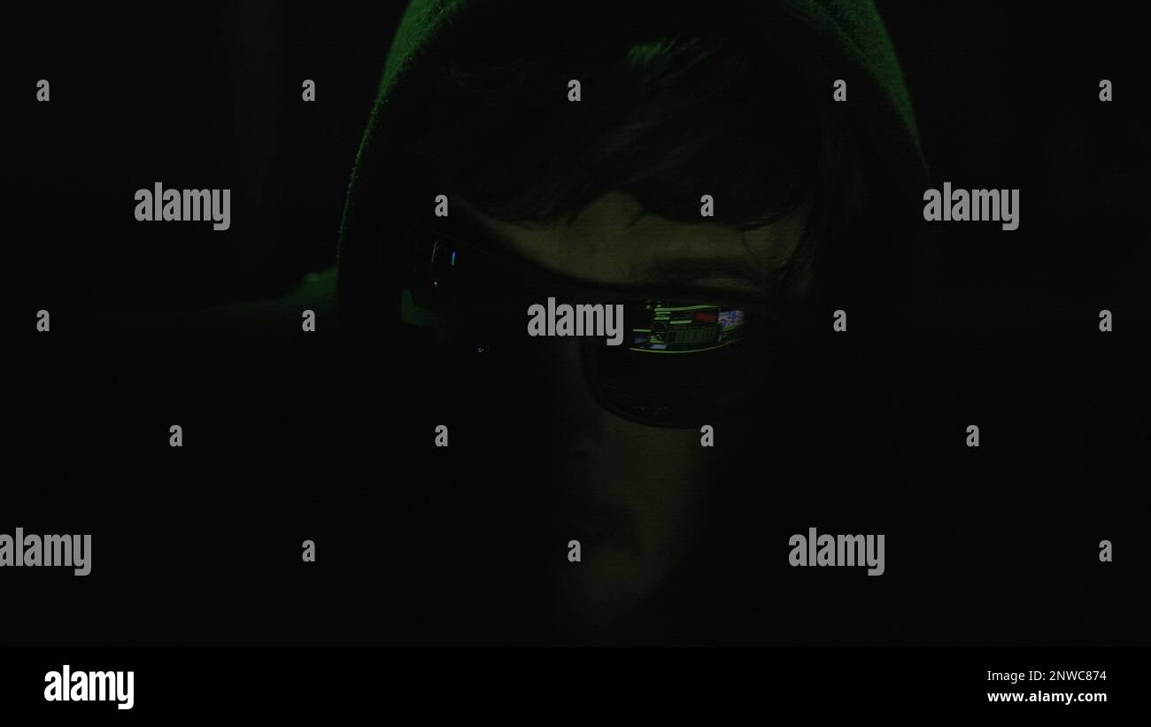 Schwarze Brille tragende Cyberkriminelle hacken in einem dunklen Raum mit geheimnisvollen grünen Lichtern, Nahaufnahme Stockfoto