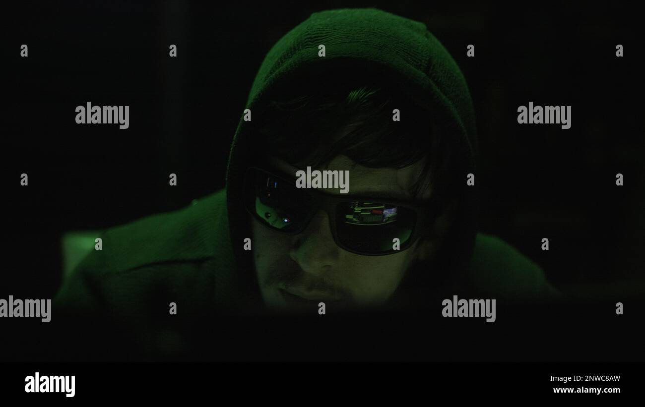 Nahaufnahme eines Hackers mit schwarzer Brille, der in einem dunklen Raum mit unheimlicher grüner Beleuchtung auf dem Computer tippt Stockfoto
