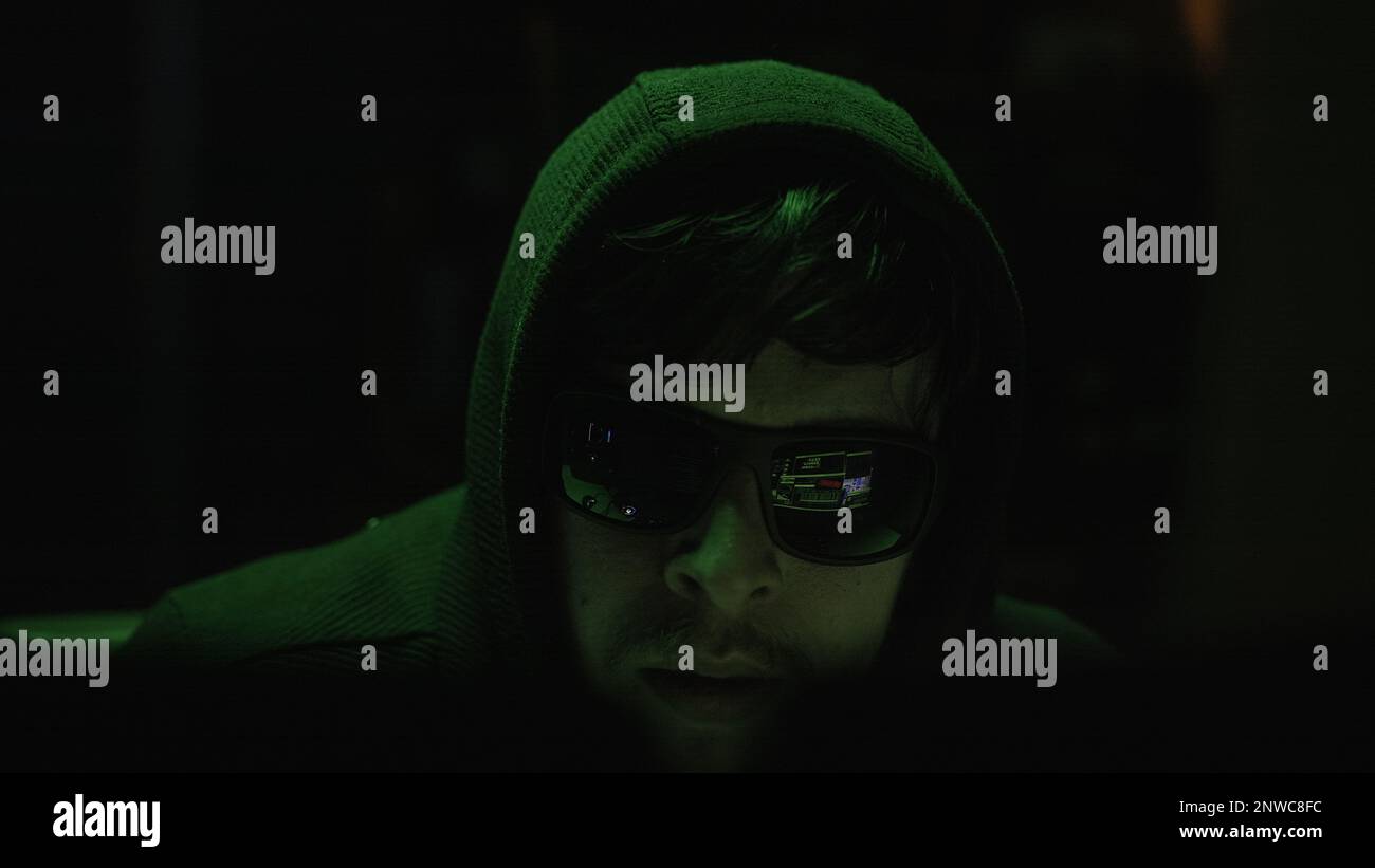Schwarze Brille tragende Cyberkriminelle hacken in einem dunklen Raum mit geheimnisvollen grünen Lichtern, Nahaufnahme Stockfoto