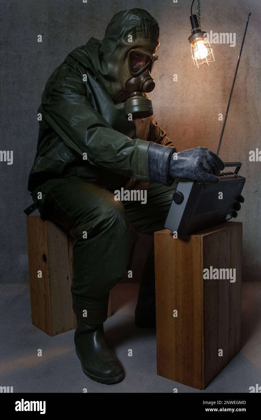 Die Rückkehr der Angst. Ein Mann in einem NBC-Schutzanzug sitzt auf Holzkisten in seinem Keller und hört sich die neuesten Nachrichten auf seinem tragbaren Radio an. Stockfoto