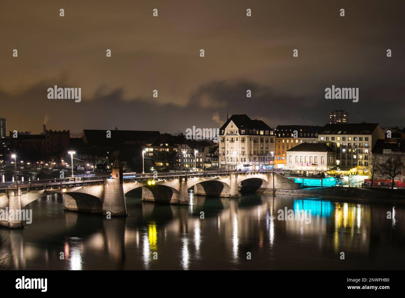 Nachtsicht auf die historische Baseler Brücke in einer rauchigen Winternacht fließt der Rhein langsam und hebt farbenfrohe Lichtreflexionen auf dem Wasser hervor Stockfoto