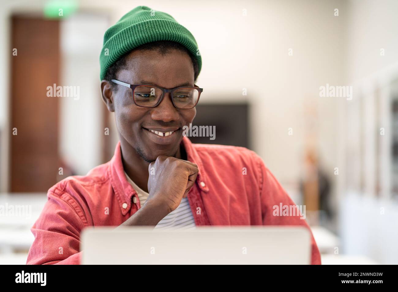Ein fröhlicher afrikanischer Student sitzt in der Bibliothek und sieht sich den Laptop-Bildschirm an und genießt E-Learning Stockfoto