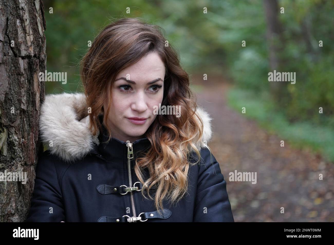 Nachdenkliche junge Frau lehnte sich gegen Baum im Wald an einem kalten Herbsttag Stockfoto