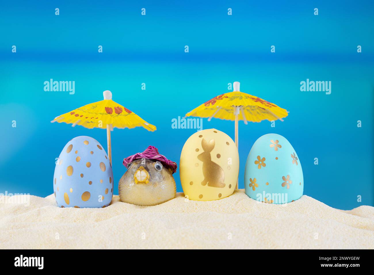 Frohe ostern. Gelbe und blaue Eier mit Kugelfisch unter Sonnenschirmen am Sandstrand des Meeres oder Ozeans. Die Feiertagspostkarte in heißem Land. Fahrt, Feder, s Stockfoto