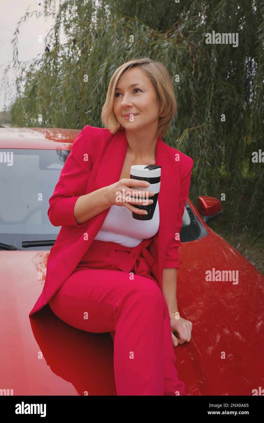 Eine Frau in einem roten chinesischen Auto trinkt Kaffee im Business-Anzug. Online-Arbeit in der Natur. Freiberuflich, unterwegs, Fernarbeit. Freizügigkeit, Modell im Wald. Hochwertiges Foto Stockfoto