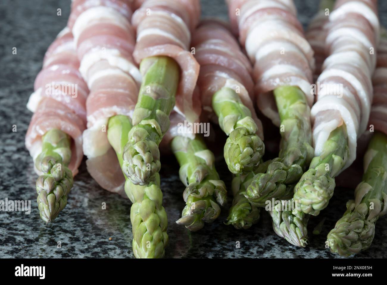 Roher organischer Spargel, in durchwachsenem Speck verpackt, auf einer schwarzen Granitarbeitsfläche. Konzept mit frischen saisonalen Produkten Stockfoto