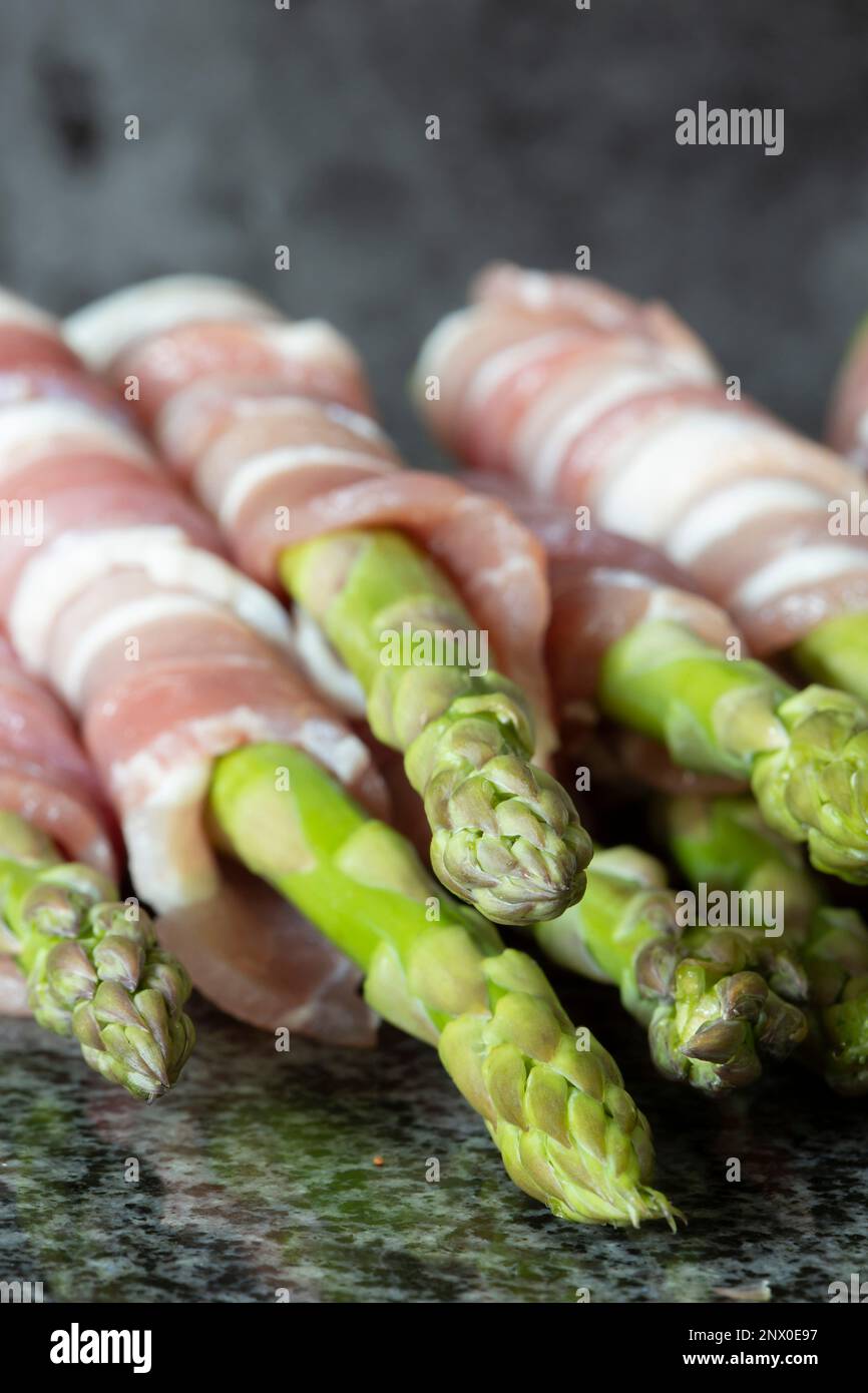 Roher organischer Spargel, in durchwachsenem Speck verpackt, auf einer schwarzen Granitarbeitsfläche. Konzept mit frischen saisonalen Produkten Stockfoto