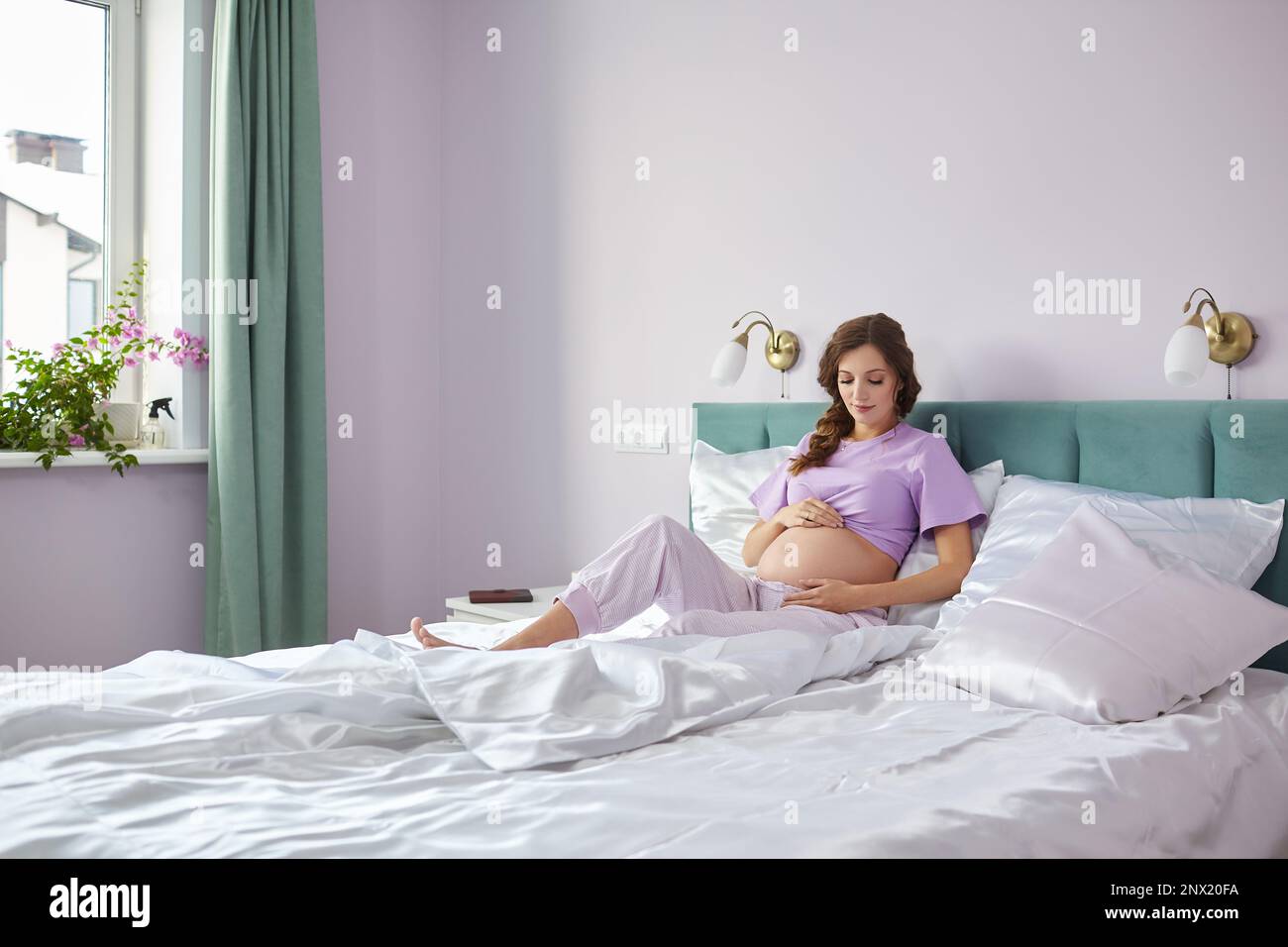 Eine schwangere Frau sitzt im Bett ihres häuslichen Schlafzimmers und schaut auf ihren Bauch. Eine Frau in Pyjama, die ihren Schwangerschaftszustand genießt. Stockfoto