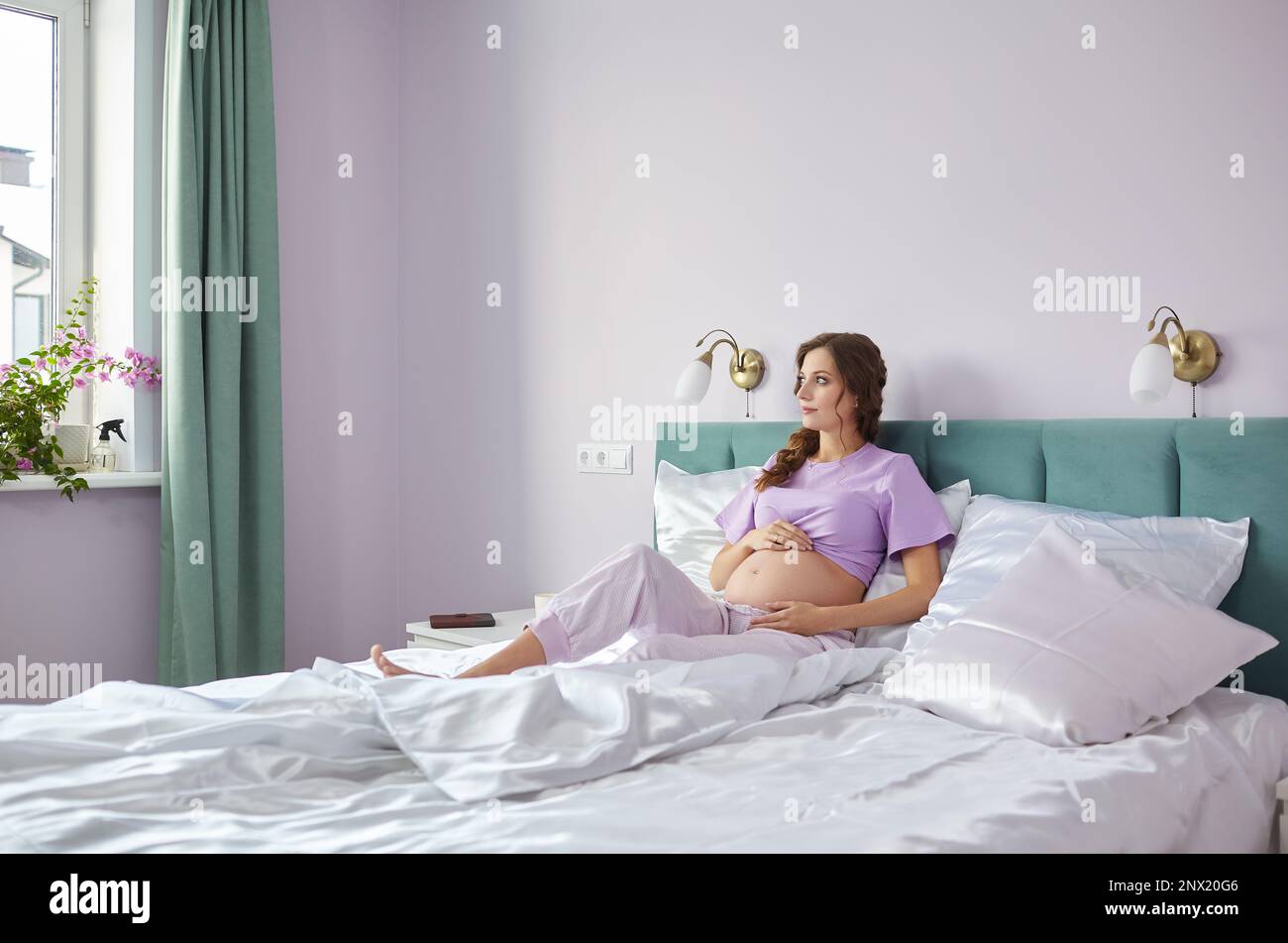 Eine schwangere Frau sitzt im Bett ihres häuslichen Schlafzimmers und schaut aus dem Fenster. Eine Frau in Pyjama, die ihren Schwangerschaftszustand genießt. Stockfoto