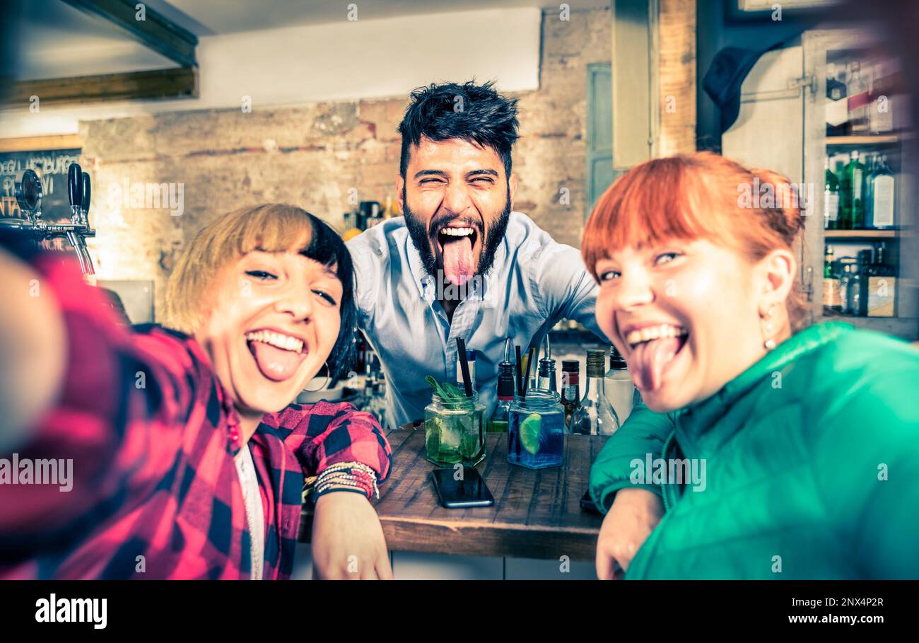 Junger gutaussehender Barkeeper flirtet mit schönen Mädchen in der Cocktailbar - glückliche Freundinnen machen Selfie mit coolem Barkeeper im Nachtclub - Vintage filt Stockfoto