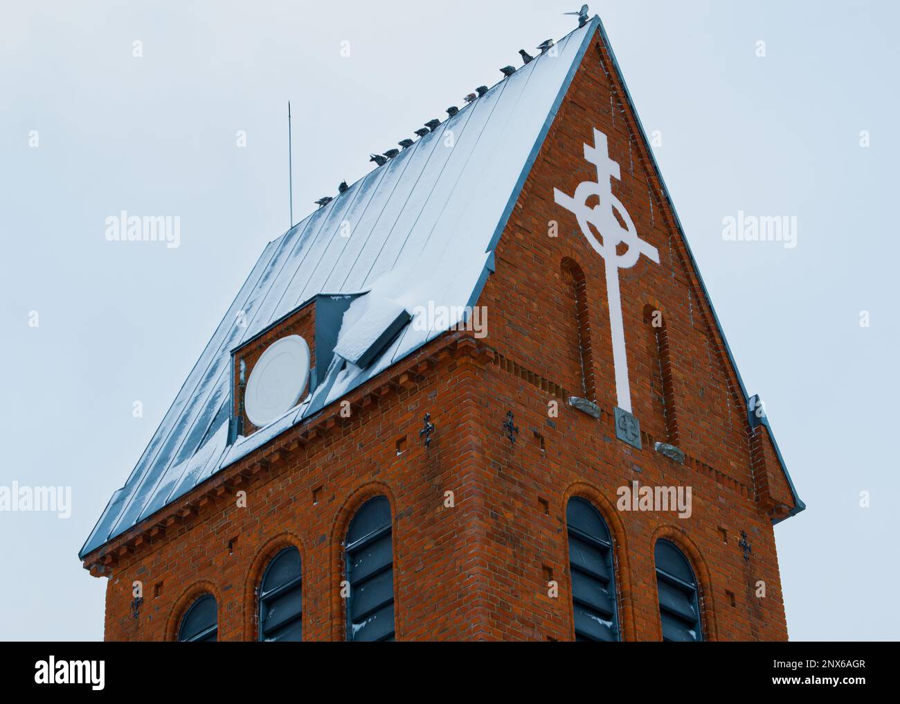 Alte rote Backsteinkirche von 1921. Im Winter fotografiert. Weißes Kreuz an der Wand. Vögel sitzen auf dem Dach. Stockfoto