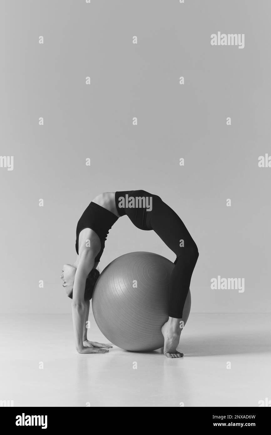 Schwarzweißfotografie. Ein junges Mädchen mit flexibler Passform, das Stretching-Übungen macht, über dem Fitnessball steht, über dem Studiohintergrund trainiert. Konzept Stockfoto
