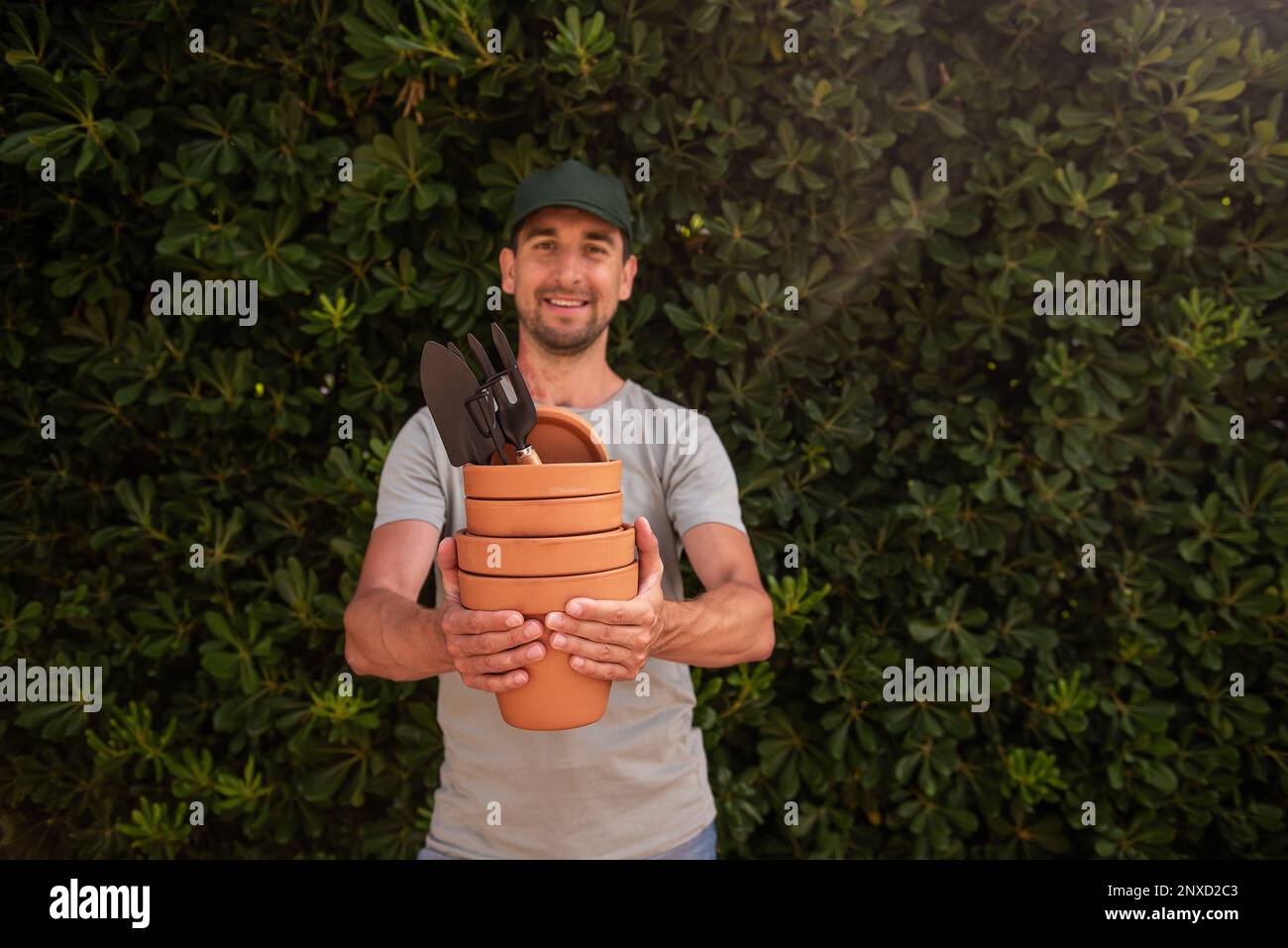 Männlicher Gärtner mit grüner Kappe hält Terrakotta-Tontöpfe mit Gartenbedarf in den Händen. Schaut in die Kamera, lächelt. Mann mittleren Alters mit roter Geburtshilfe Stockfoto