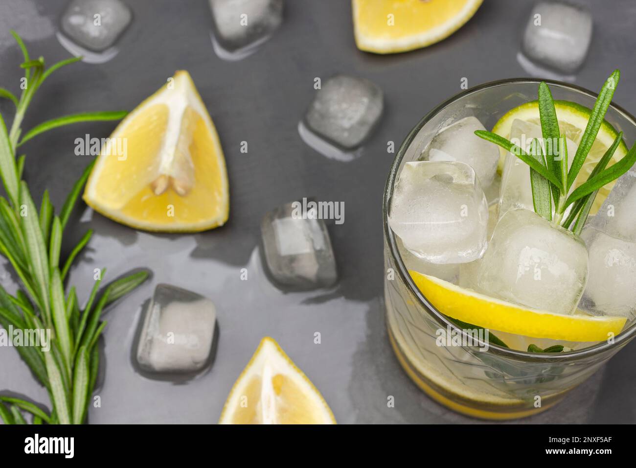 Zitronen-, Eis- und Rosmarin-Zweig in einem Glas. Geschmolzenes Eis auf dem Tisch. Flach verlegt. Grauer Hintergrund. Stockfoto