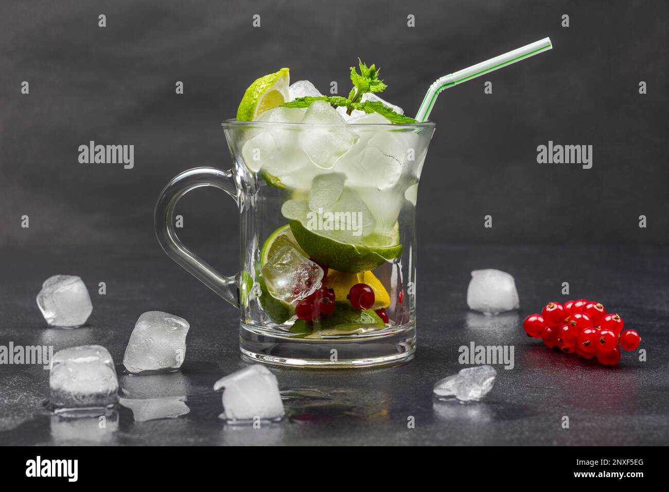 Limette, rote Johannisbeere, Minze, Eis und Strohhalme in einem Glas. Geschmolzenes Eis und ein Zweig rote Johannisbeere auf dem Tisch. Schwarzer Hintergrund. Stockfoto