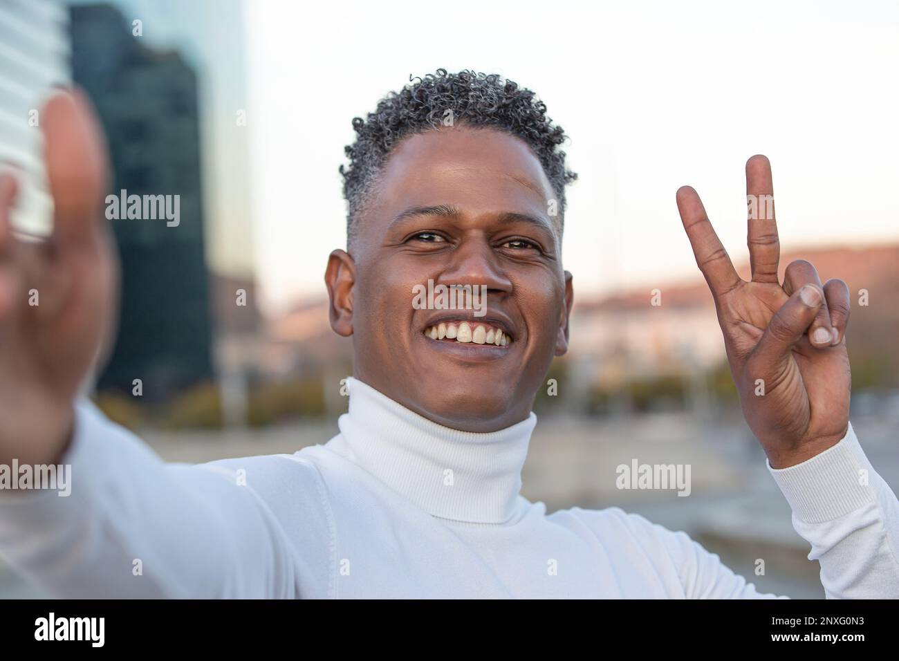 Lächelnder junger Mann, der Siegesgeste zeigt, während er Selfie in der Stadt macht Stockfoto