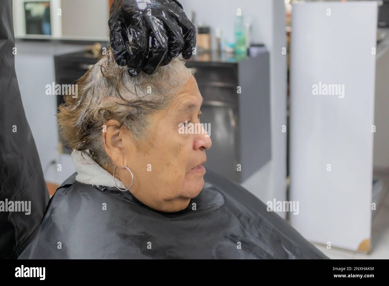 Friseurin massiert kurze Haare älterer Frauen mit Farbstoff vor verschwommenem Hintergrund, ernster Ausdruck, Seitenansicht. Alte Mode und Schönheitskonzert Stockfoto
