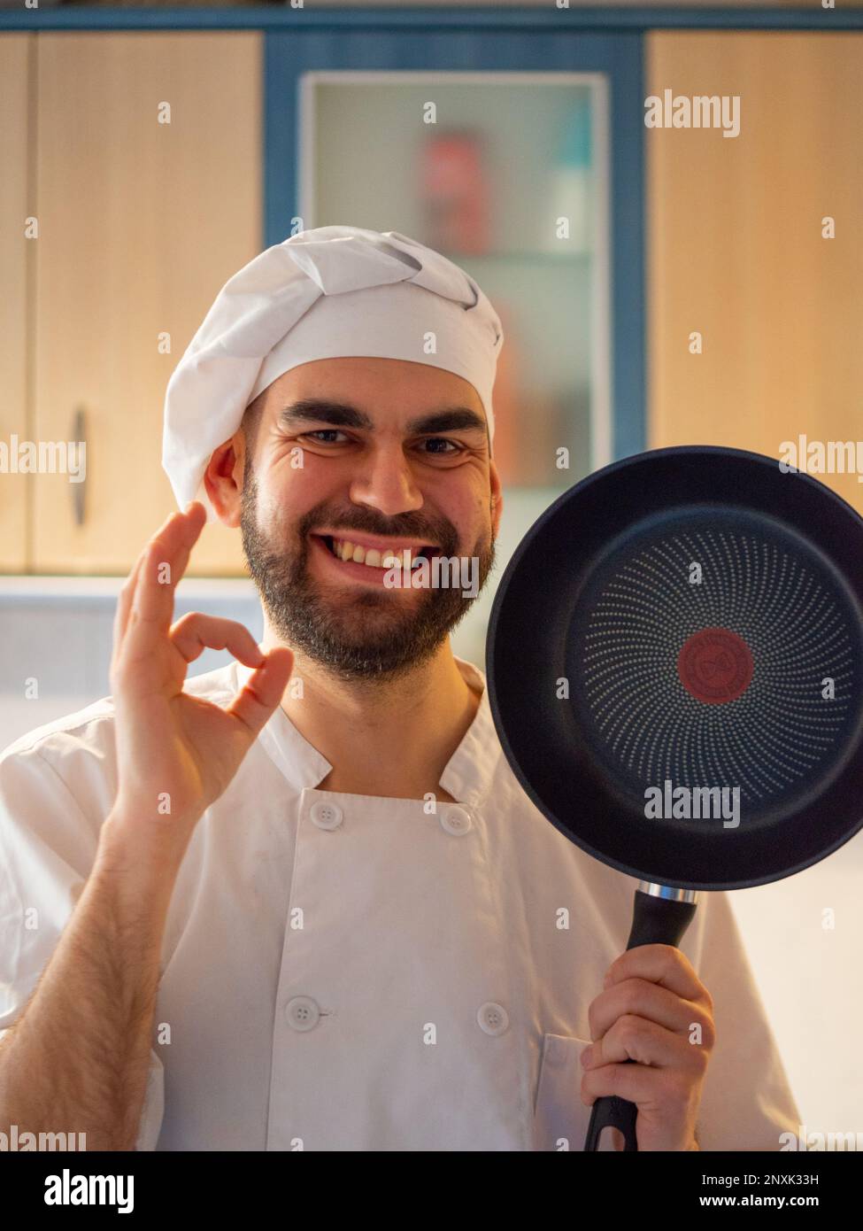 Porträt eines jungen, bärtigen Kochs, der lächelt, während er eine Antihaftpfanne zeigt, die in die Kamera schaut Stockfoto