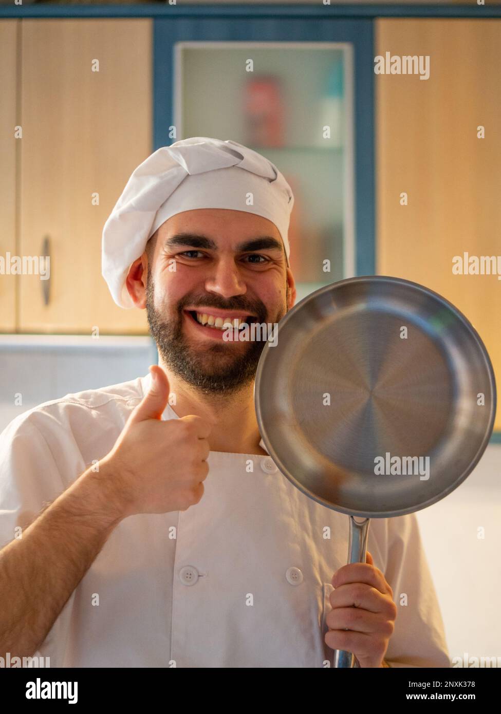 Porträt eines jungen, bärtigen Kochs, der lächelt, während er mit einer Edelstahlpfanne in die Kamera schaut Stockfoto