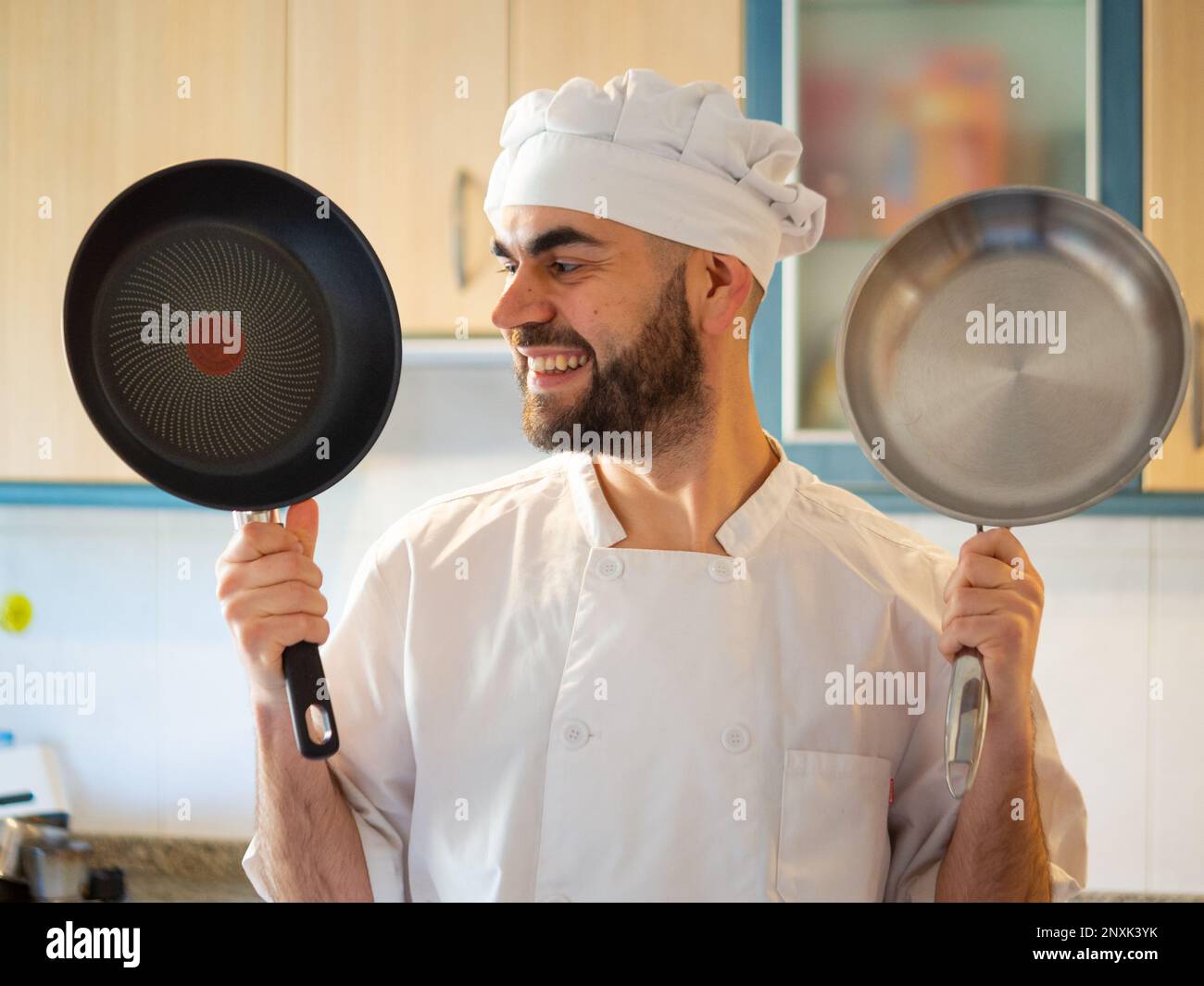 Porträt eines jungen, bärtigen Chefkochs, der lächelt, während er eine antihaftbeschichtete Pfanne und eine Edelstahlpfanne zeigt Stockfoto