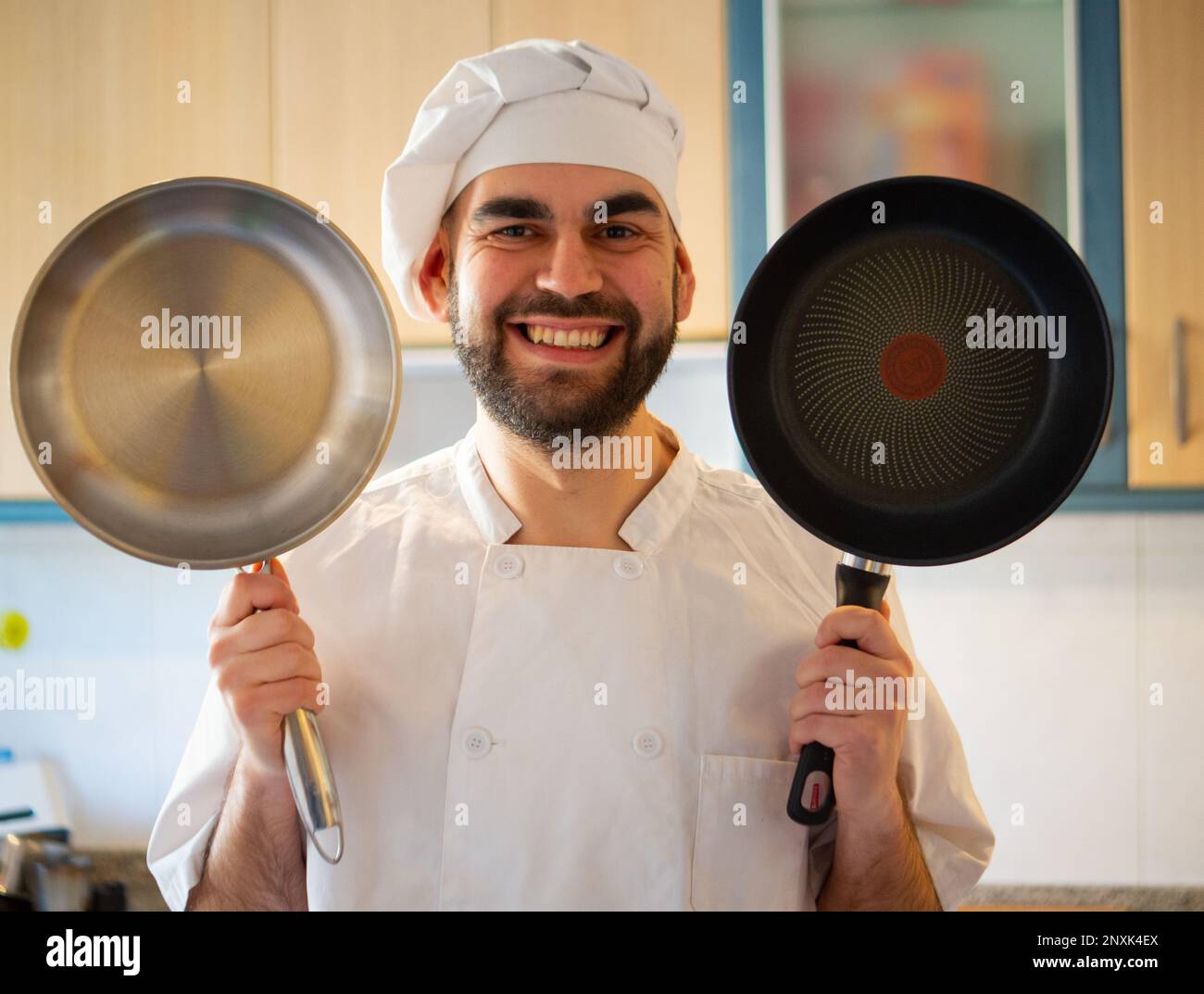 Porträt eines jungen, bärtigen Kochs, der lächelt, während er eine antihaftbeschichtete Pfanne und eine Edelstahlpfanne in die Kamera zeigt Stockfoto