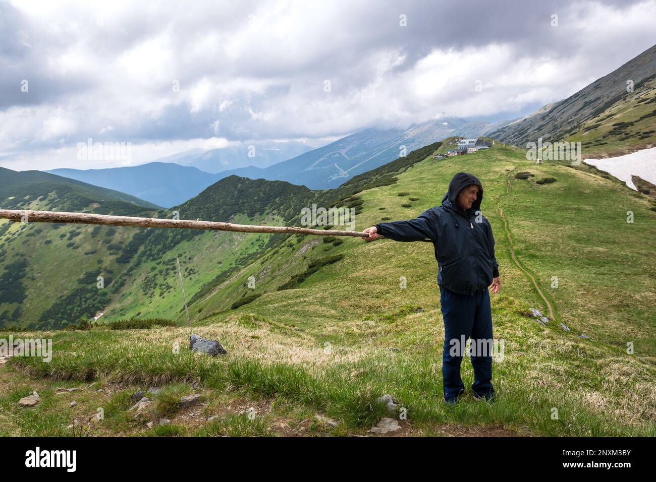 Ein Mann hält eine Holzstange in der Hand auf einem der Gipfel der niedrigen Tatra, mit dem Štefánika Holiday House im Hintergrund, Brezno, Slowakei Stockfoto