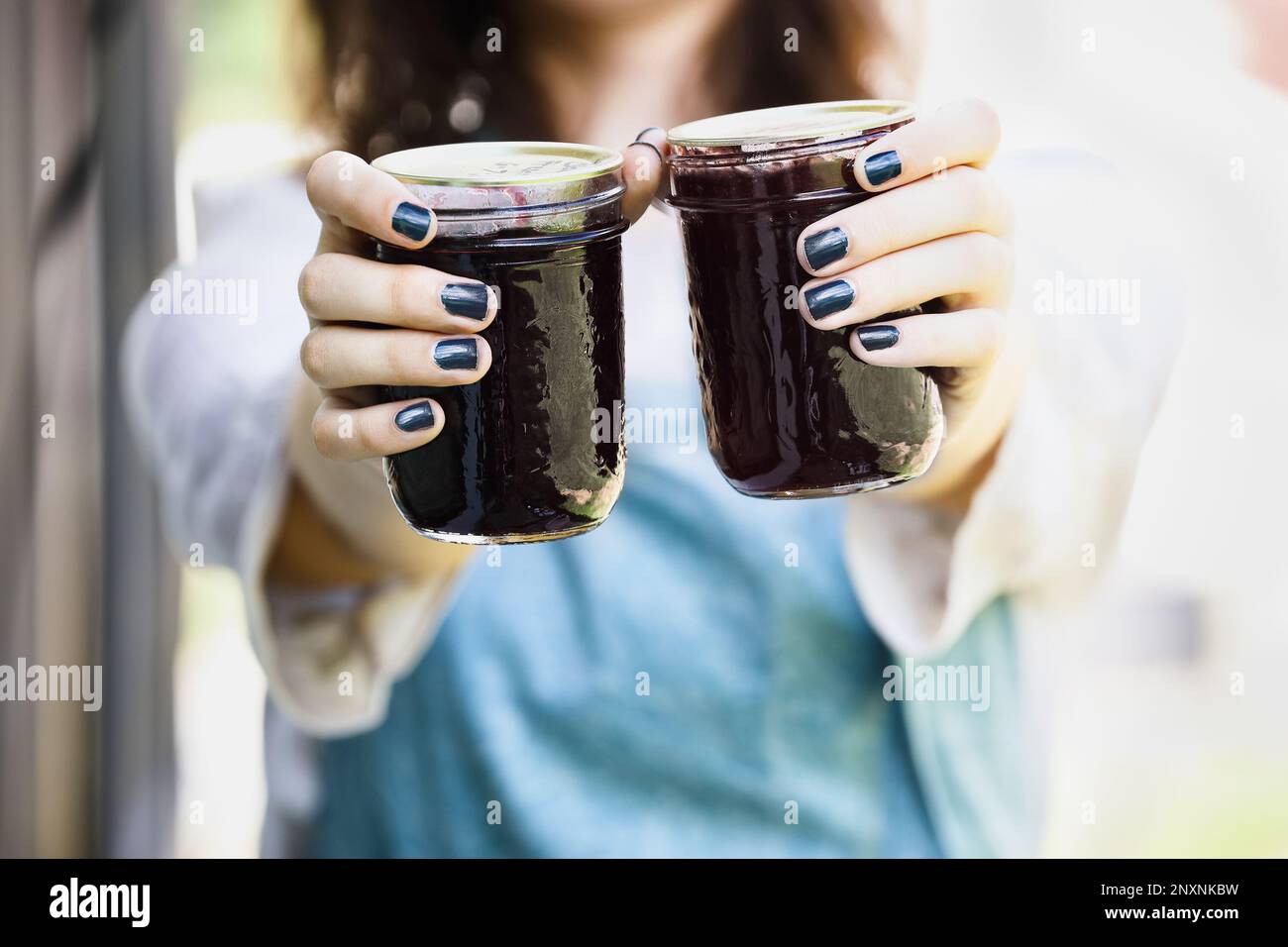 Eine Frau in blauer Schürze, die hausgemachte Brombeermarmelade in der Hand hält. Selektiver Fokus auf Maurerglas mit unscharfem Hintergrund. Stockfoto