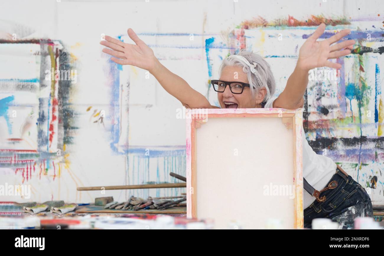 Ältere grauhaarige, reife, fröhliche Künstlerin mit Brille hinter Leinwand streckt ihre Arme mit Kraft und Freude in ihrem Studio, Copy Spac Stockfoto
