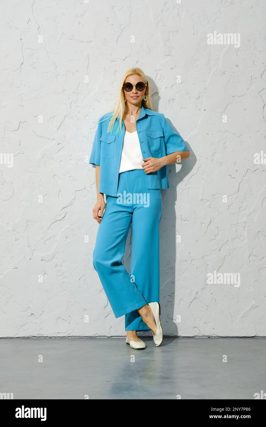 Modemodell in Sonnenbrille in blauer Jacke und Hose, die sich gegen die strukturierte weiße Wand lehnen Stockfoto