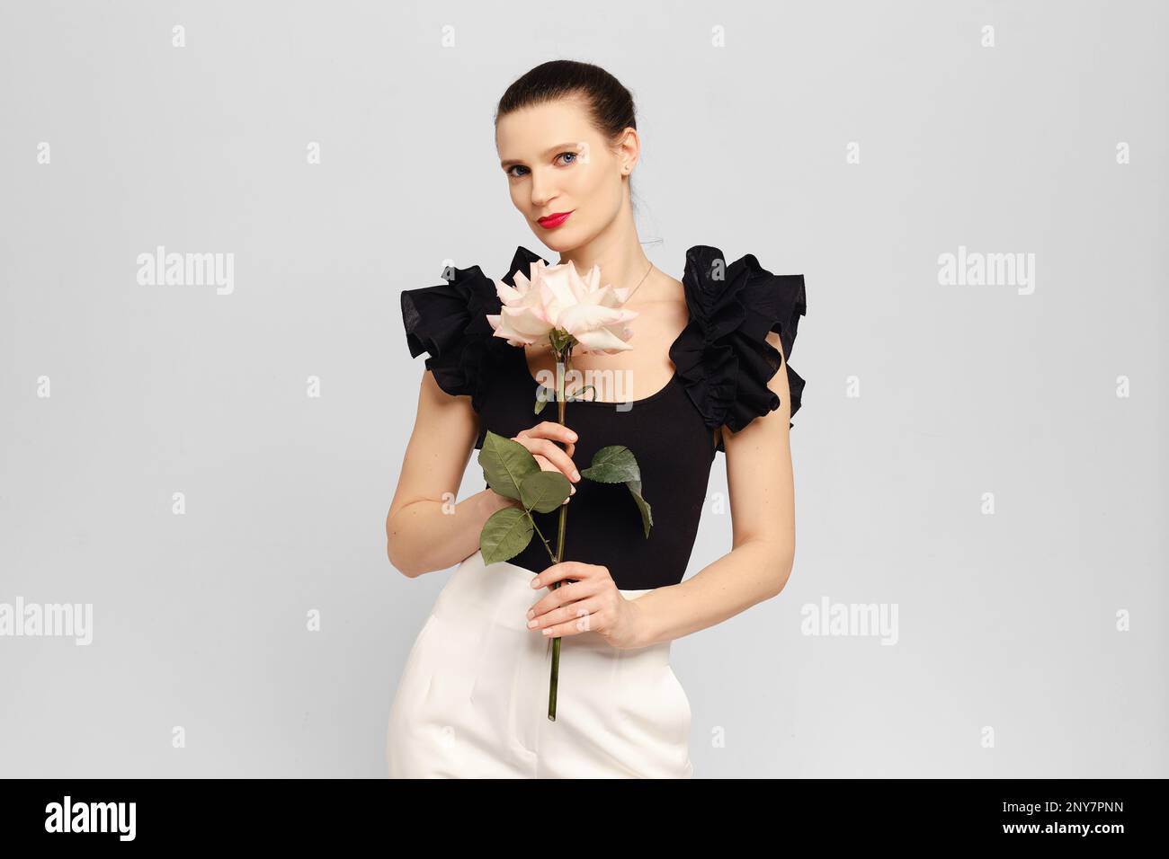 Stilvolle und positive Erwachsene Frau, die im weißen Studio posiert und Rose in den Händen hat Stockfoto