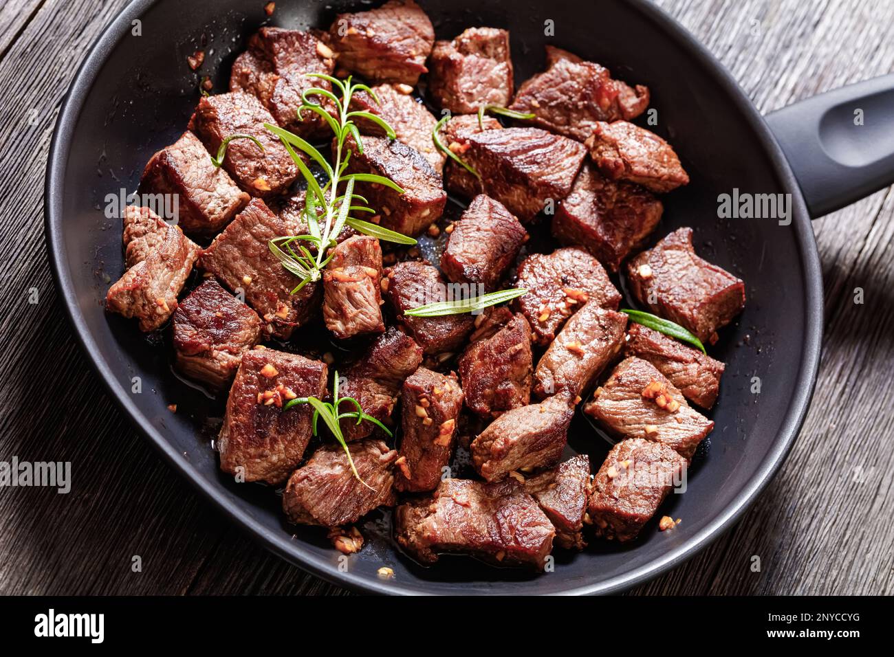 Gebratene saftige New yorker Steak-Bissen mit Butter, Rosmarin und Knoblauch auf Pfannkuchen auf einem dunklen Holztisch, Nahaufnahme Stockfoto