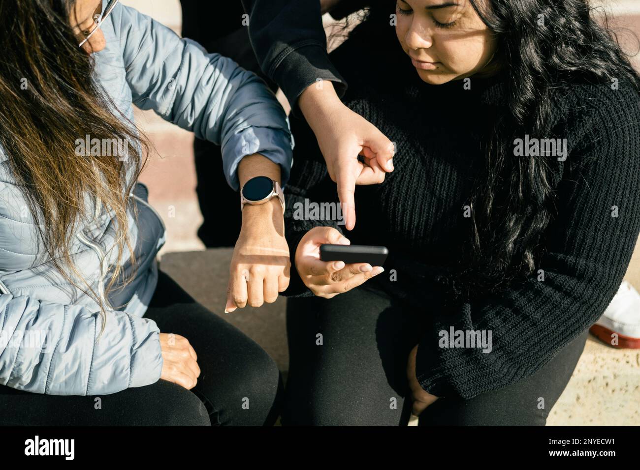 Glückliche Freunde teilen Multimedia-Inhalte online mit ihren Smartphones und ihrer Smartwatch, zwei wunderschöne Latina-Frauen, die draußen sitzen Stockfoto