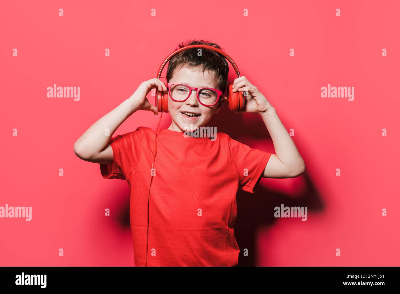 Positiver Junge in rotem T-Shirt und Brille, der Kopfhörer berührt, während er im Studio den Lieblingssong hört und wegschaut Stockfoto