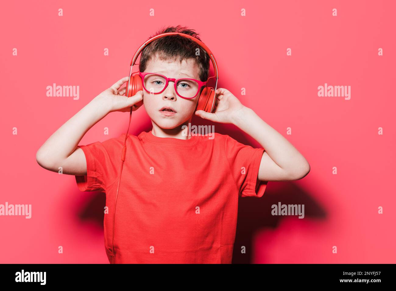 Positiver Junge in rotem T-Shirt und Brille, der Kopfhörer berührt, während er im Studio den Lieblingssong hört und in die Kamera schaut Stockfoto
