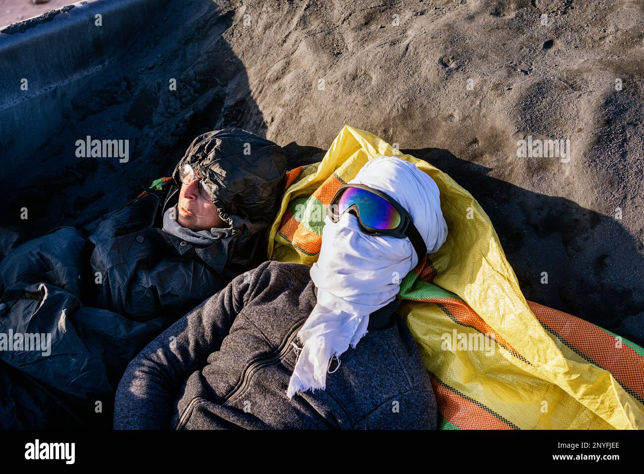 Von oben: Muslimischer Mann mit Gesichtsschutz und Schutzbrille, die auf sandigem Boden in der Nähe einer Frau in warmer Jacke mit Kapuze auf dem Kopf liegt Stockfoto