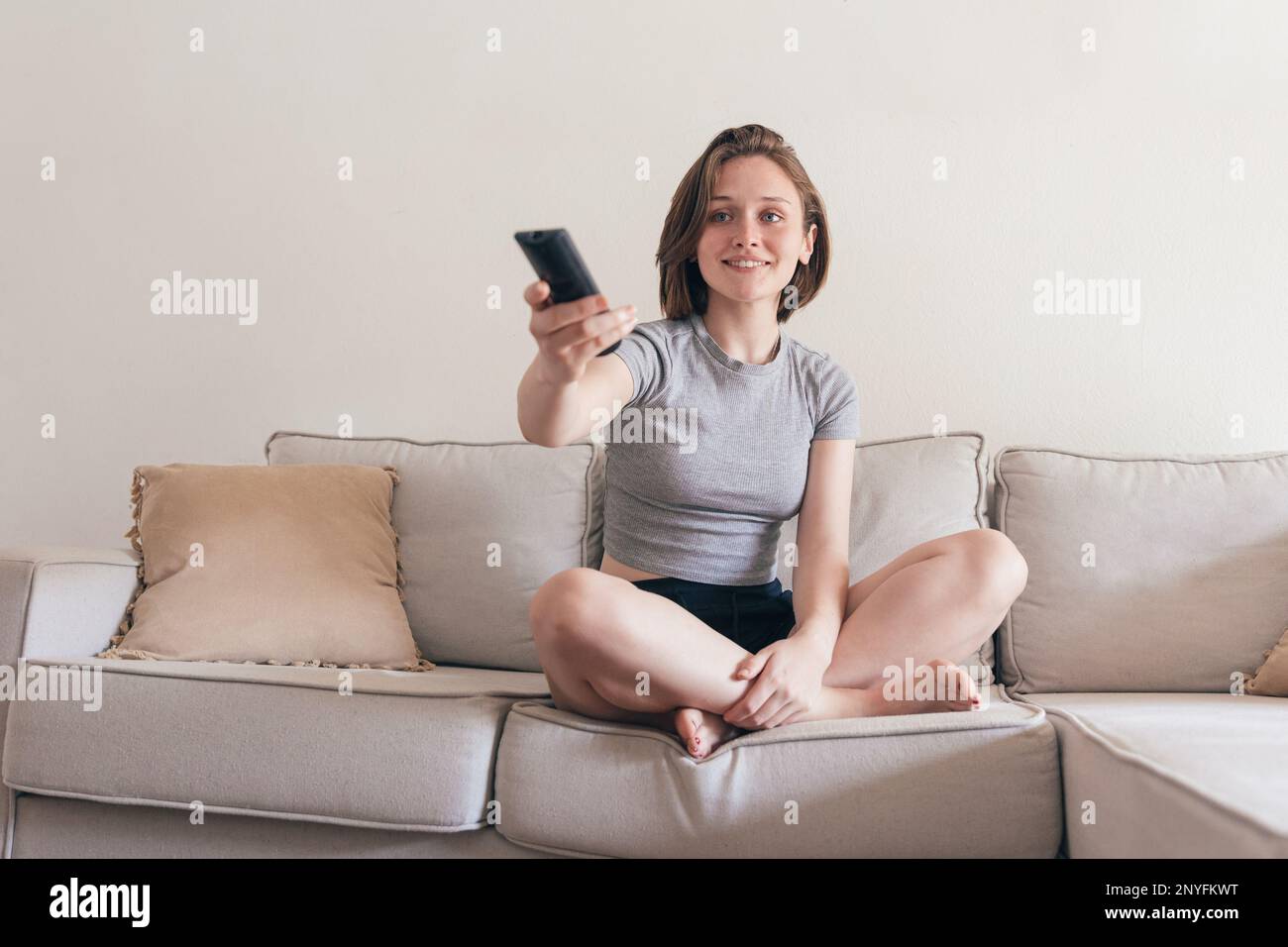 Voller Körper einer positiven jungen Frau in Haushaltskleidung, die auf dem Sofa sitzt, sich die Beine kreuzte und lächelte, während sie mit der Fernbedienung A den Kanal wechselte Stockfoto