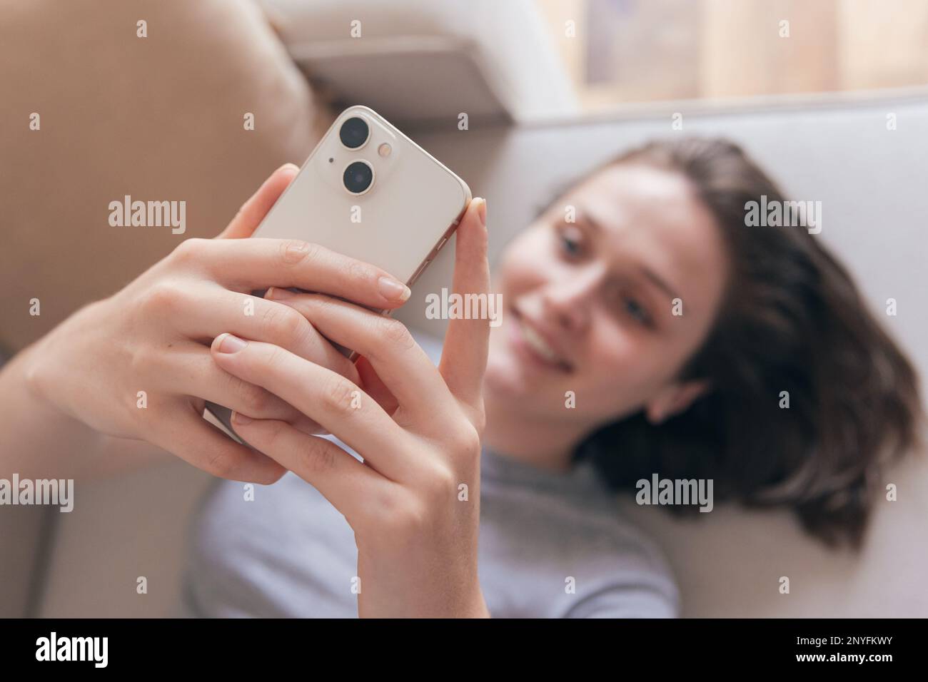 Von oben eine glückliche junge Frau im grauen T-Shirt lächelt und surft im Internet auf dem Handy, während sie zu Hause auf dem Sofa liegt Stockfoto