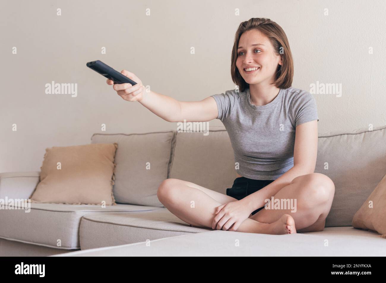 Voller Körper einer positiven jungen Frau in Haushaltskleidung, die auf dem Sofa sitzt, sich die Beine kreuzte und lächelte, während sie mit der Fernbedienung A den Kanal wechselte Stockfoto