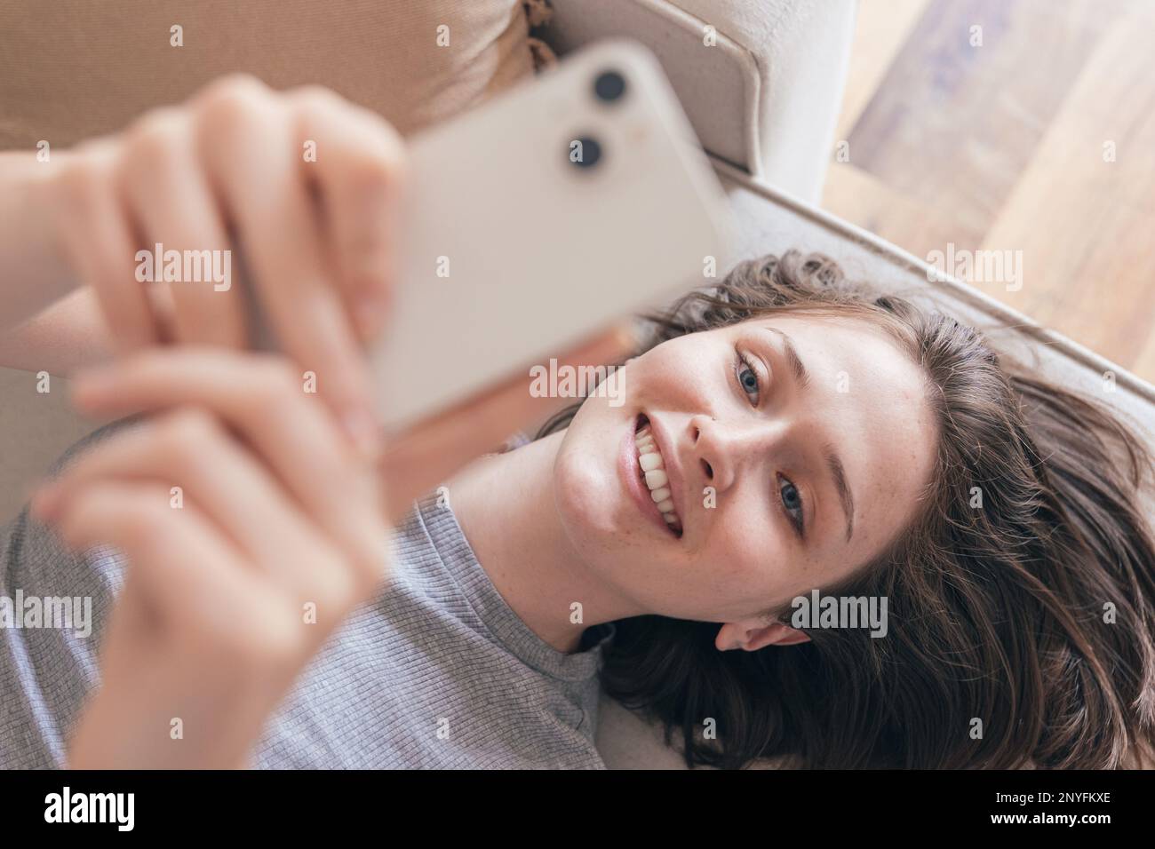 Von oben eine glückliche junge Frau im grauen T-Shirt lächelt und surft im Internet auf dem Handy, während sie zu Hause auf dem Sofa liegt Stockfoto