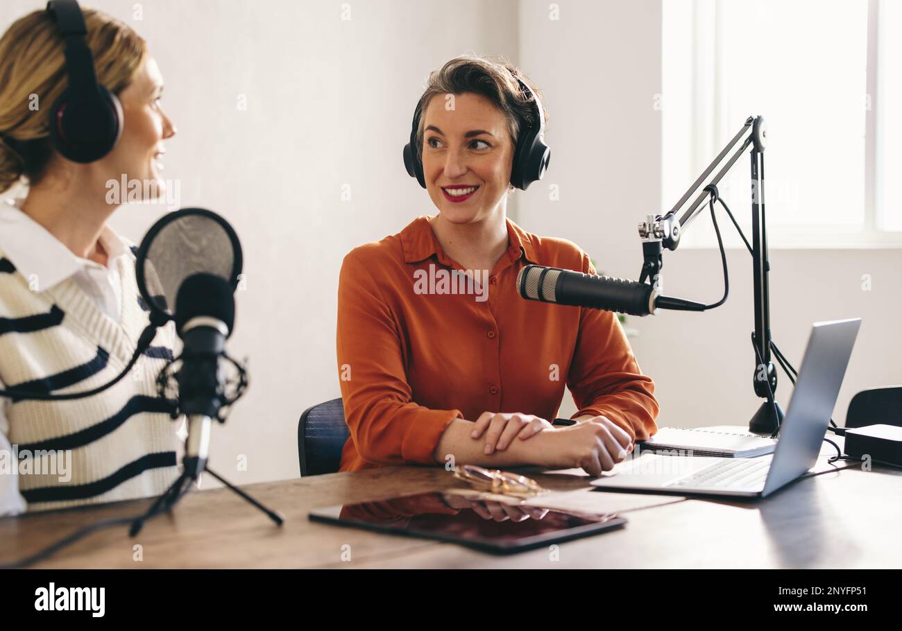 Zwei Frauen, die sich unterhalten, während sie eine Audioübertragung in einem Heimstudio moderieren. Zwei weibliche Content-Ersteller nehmen einen Internet-Podcast auf Stockfoto
