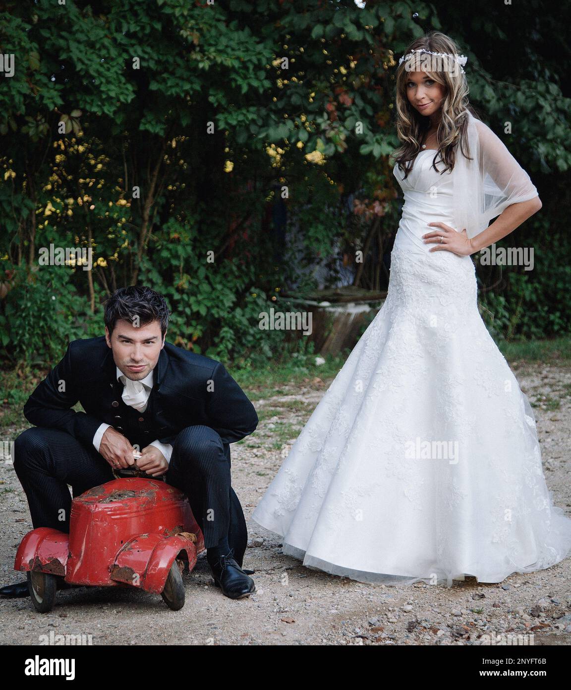 Attraktive junge Braut und Bräutigam lächeln beim Spielen mit alten Spielzeugauto im Garten Stockfoto
