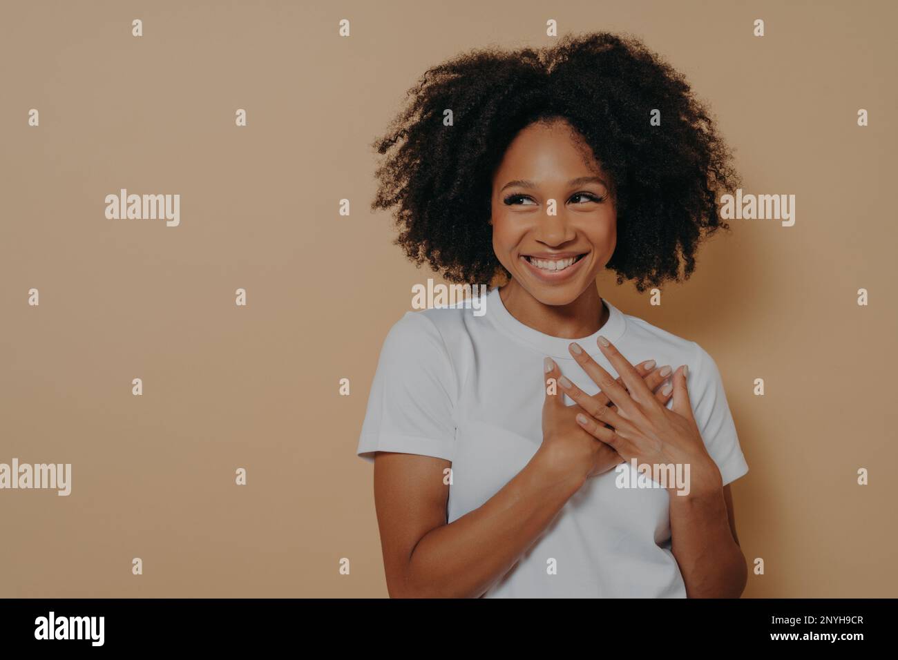 Porträt einer glücklichen afrikanerin in einem weißen T-Shirt, das auf beigefarbenem Hintergrund posiert, mit einem Lächeln, das wegschaut, Hände auf der Brust hält, fühlt sich Dankbarkeit und Geste an Stockfoto