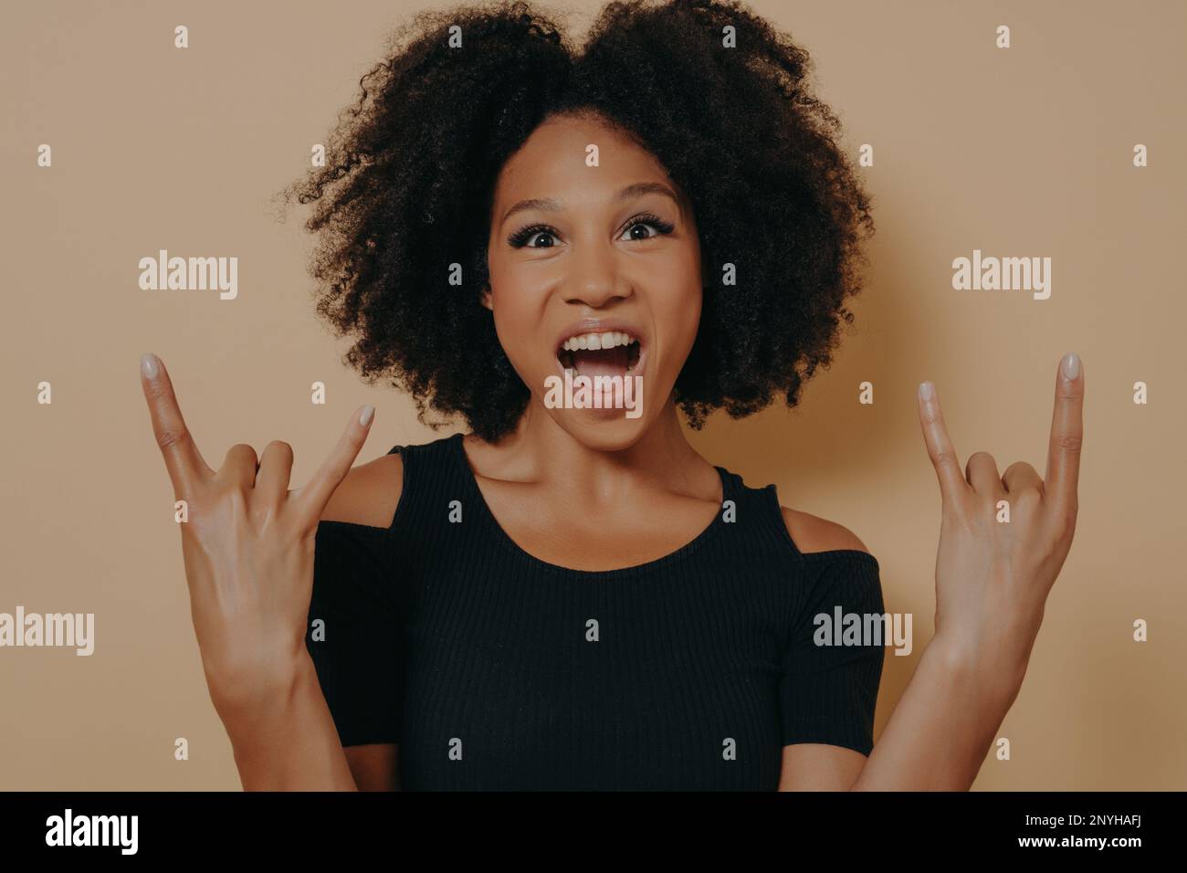 Junge afroamerikanische Frau, die ein schwarzes T-Shirt trägt, mit verrücktem Gesichtsausdruck schreit und Rock-n-Roll-Symbol macht, mit erhobenen Händen wie der Musikstar Isolat Stockfoto