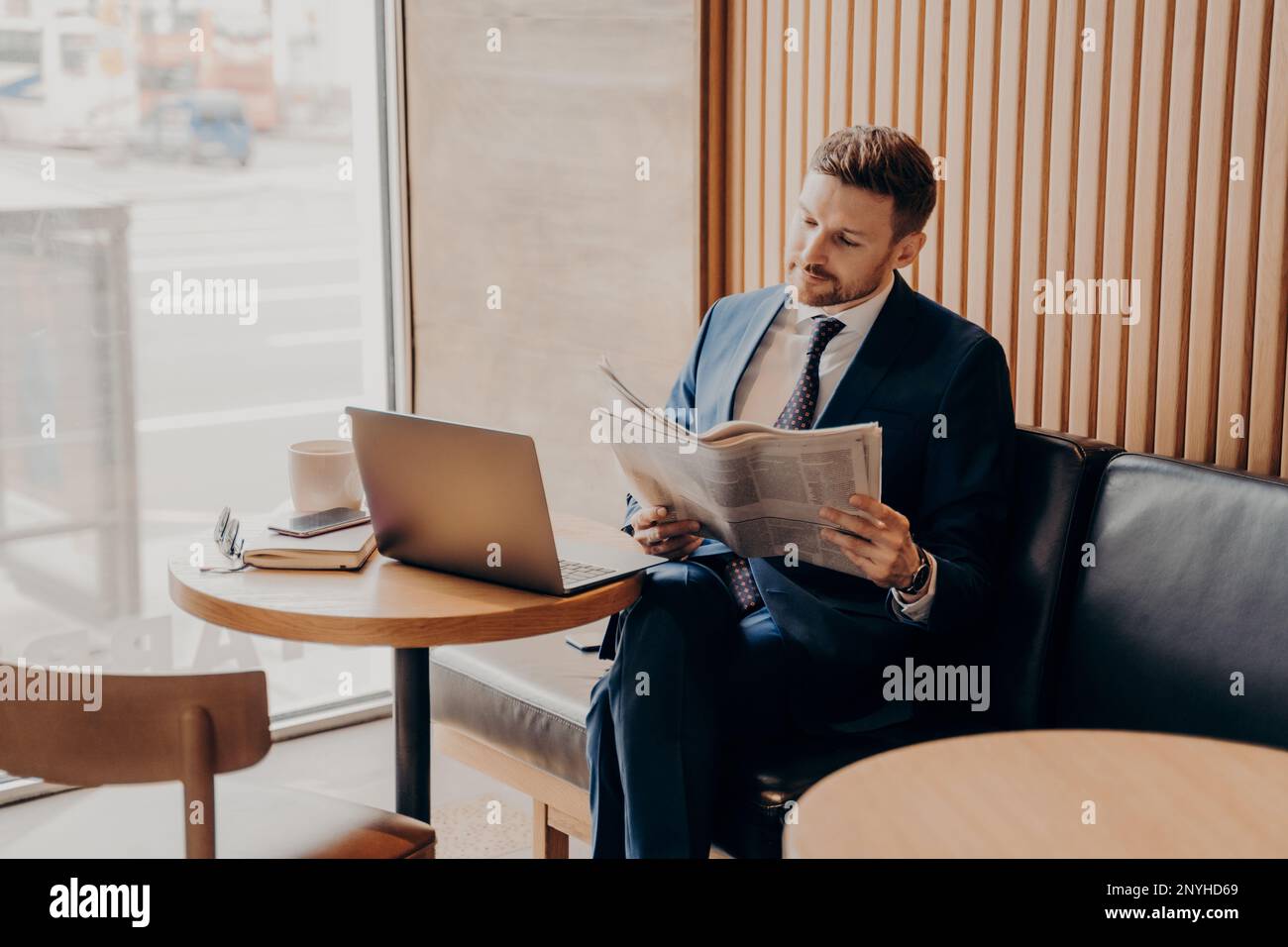 Fokussierter, reicher junger Geschäftsmann in blauem Anzug und weißem Hemd liest Nachrichten über sein Startup-Unternehmen in einer lokalen Zeitung, während er allein im gemütlichen Café sitzt Stockfoto