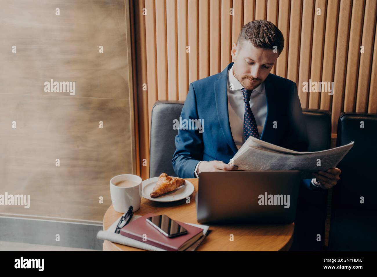 Junger, knackiger Unternehmer in formeller Kleidung, der sich während der Kaffeepause auf die Lektüre von Finanzzeitungen konzentrierte, während er seinen Cappuccino an genoss Stockfoto